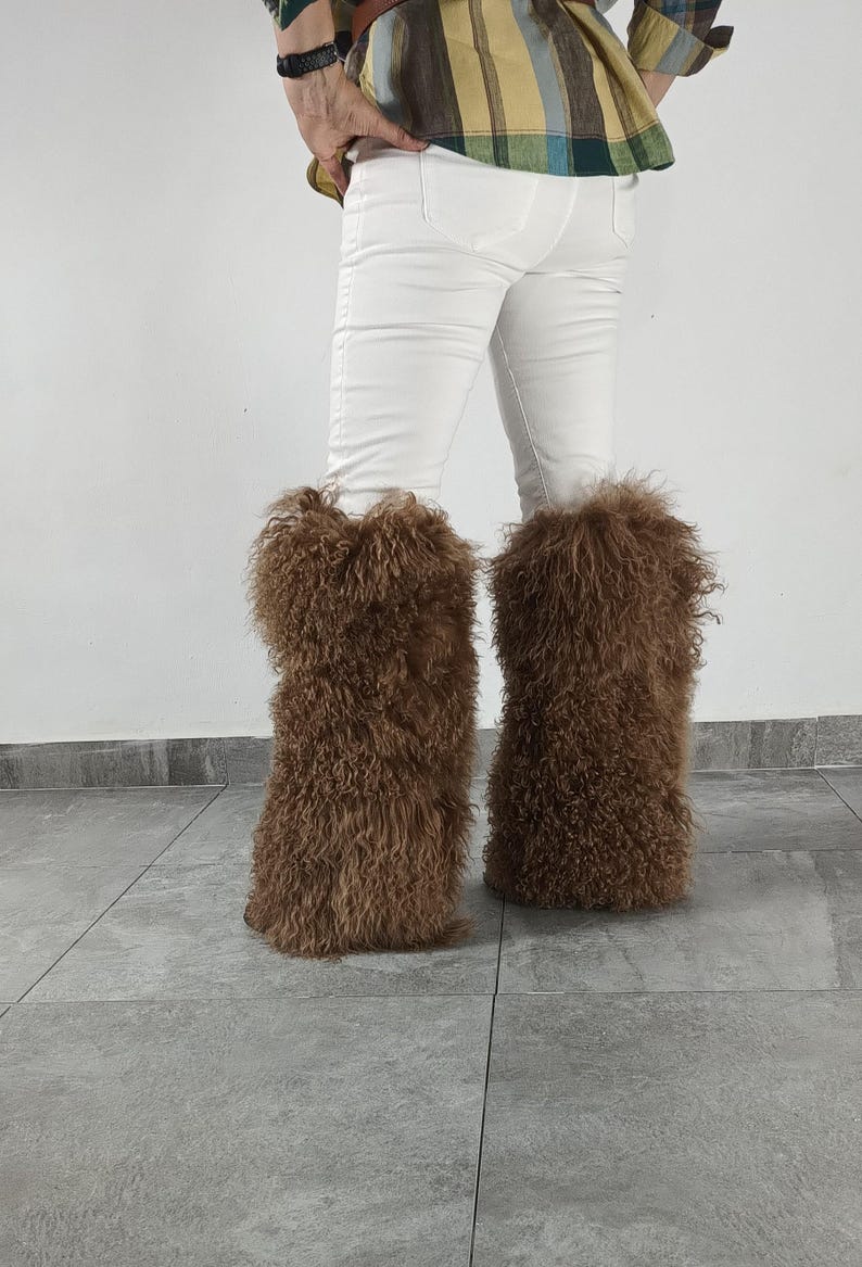 May include: A pair of brown, long-haired, shaggy faux fur leg warmers worn over white pants. The leg warmers feature a dense, curly texture that extends from the lower calf to the ankle, providing a voluminous appearance around the lower legs.