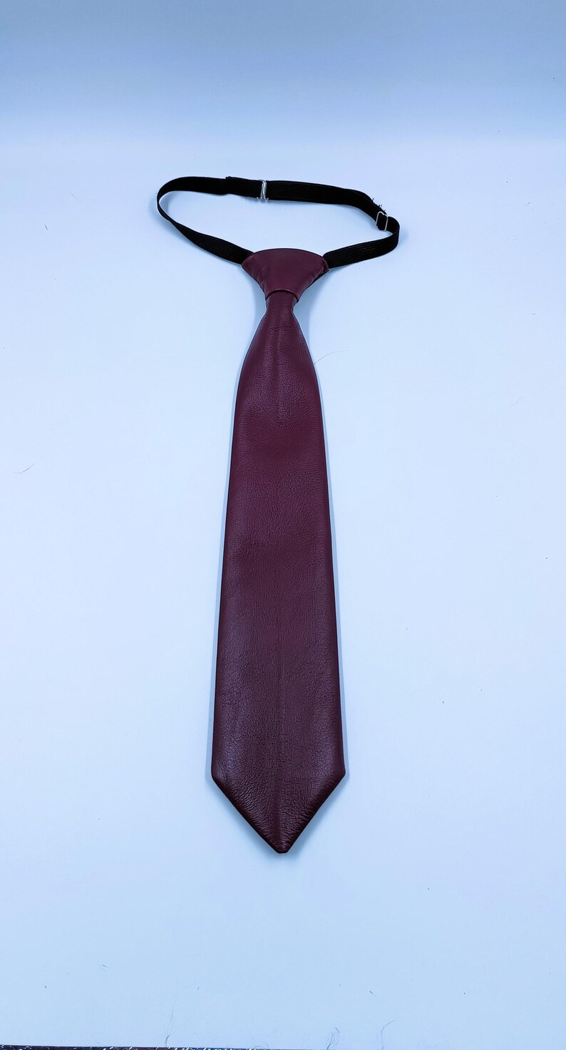 Peut inclure: Une cravate bordeaux profond fabriqu&eacute;e dans une mati&egrave;re textur&eacute;e au grain visible. La cravate pr&eacute;sente une extr&eacute;mit&eacute; pointue et un n&oelig;ud pr&eacute;-nou&eacute; attach&eacute; &agrave; une sangle de cou &eacute;lastique noire avec un curseur m&eacute;tallique r&eacute;glable pour un ajustement s&ucirc;r.