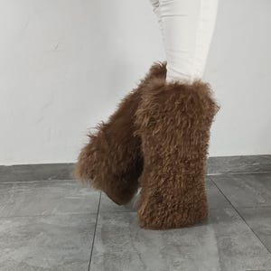 May include: A pair of tall, brown, shaggy faux fur leg warmers worn over white pants. The leg warmers feature a long, textured, curly pile that covers the lower legs, extending from the mid-calf down to the ankles, creating a voluminous appearance.
