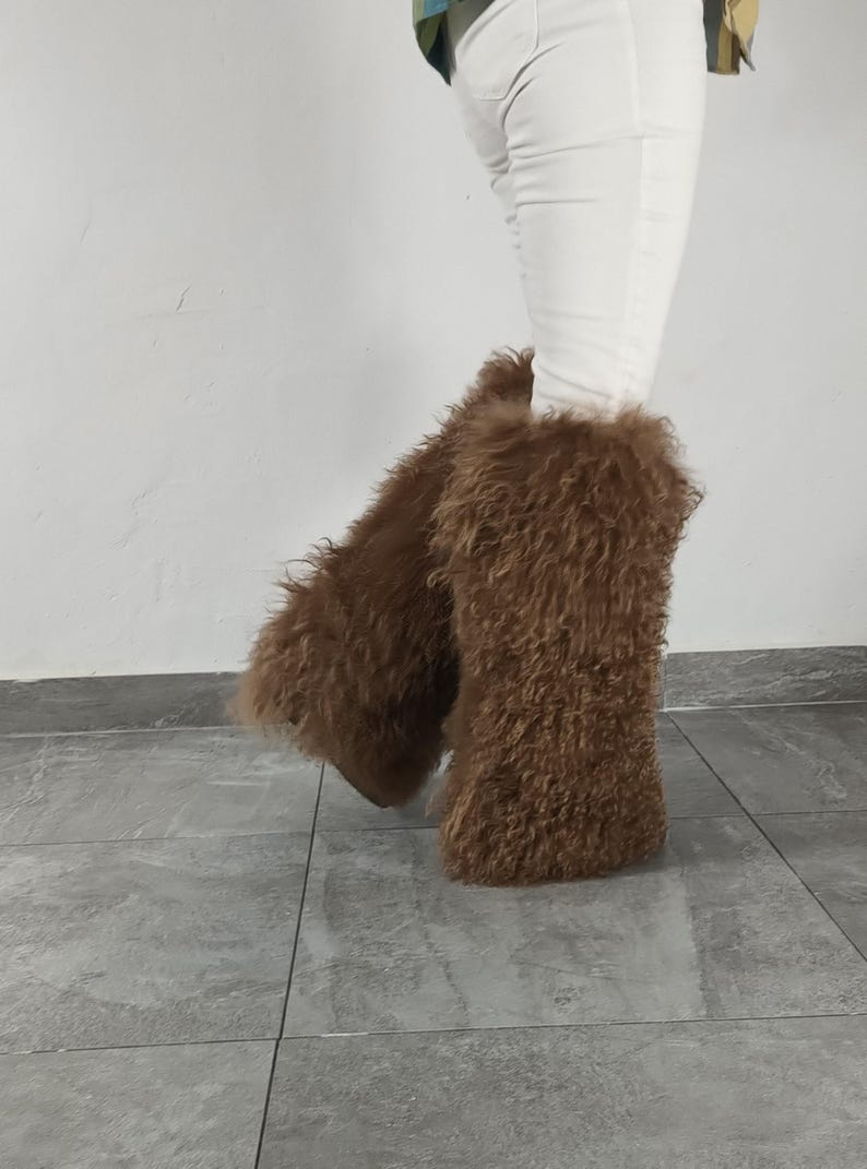 May include: A pair of tall, brown, shaggy faux fur leg warmers worn over white pants. The leg warmers feature a long, textured, curly pile that covers the lower legs, extending from the mid-calf down to the ankles, creating a voluminous appearance.