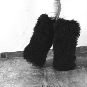 May include: A pair of black, long, shaggy faux fur leg warmers worn over light-colored pants. The leg warmers feature a dense, textured, and voluminous pile that extends from the mid-calf down to the ankles, creating a full and fuzzy appearance.