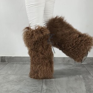 May include: A pair of brown, long-haired, fluffy fur leg warmers worn over white pants. The leg warmers feature a textured, curly appearance and include a small black logo patch with the text Z FURS visible on the front of the left leg warmer.