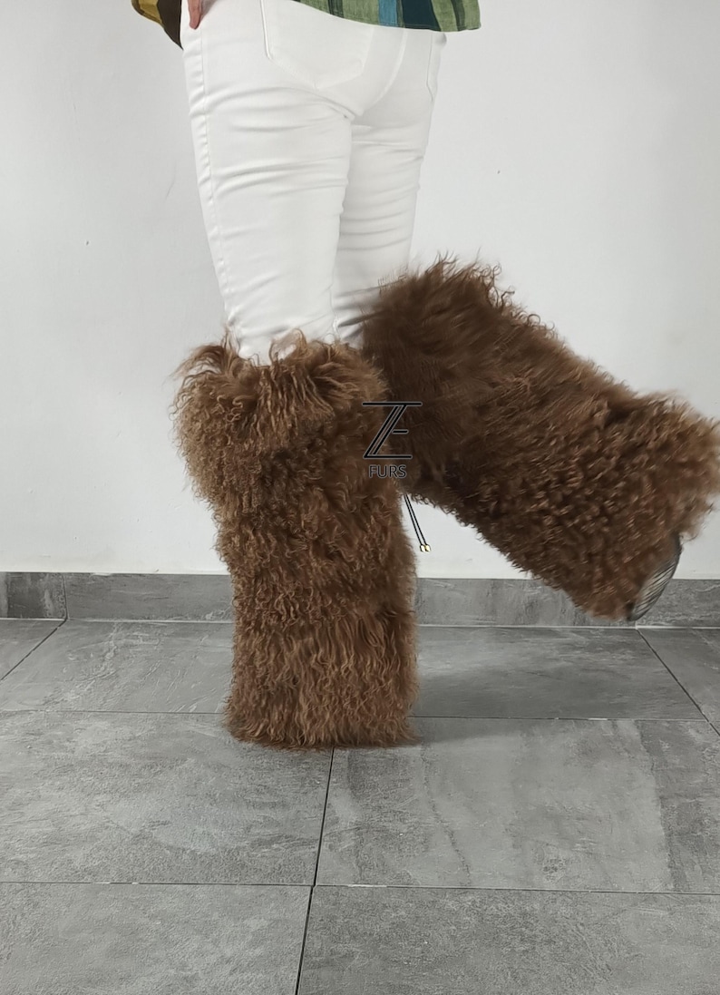 May include: A pair of brown, long-haired, fluffy fur leg warmers worn over white pants. The leg warmers feature a textured, curly appearance and include a small black logo patch with the text Z FURS visible on the front of the left leg warmer.