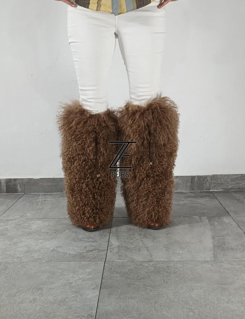 May include: A pair of tall, brown, curly-haired fur leg warmers worn over white pants. The leg warmers feature a thick, textured, shaggy appearance and are adorned with a black logo featuring the letter Z and the text FURS in the center.