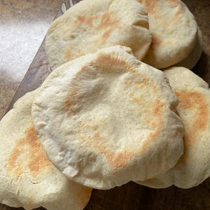 Puede incluir: Varios panes de pita recién horneados, de color claro con manchas doradas. Los panes planos redondos están dispuestos sobre una tabla de madera, destacando su textura suave y su aspecto casero.