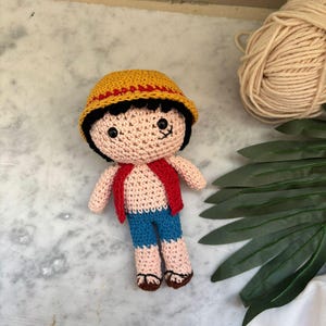 Könnte beinhalten: Handgefertigte Häkelpuppe mit gelbem Hut und rotem Band, schwarzem Haar und roter Weste. Die Puppe hat blaue Shorts, gebräunte Haut und braune Schuhe. Ein einzigartiger, handgefertigter Artikel.