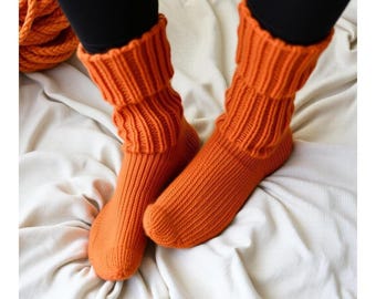 May include: A pair of orange knitted socks with a ribbed pattern. The socks are folded down at the top, showing the ribbed texture. The socks are on a white surface.