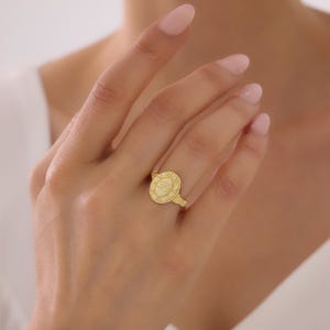 Op de afbeelding: Een gouden zegelring met een ovaalvormig vlak dat in het midden is gegraveerd met een cursieve letter L. Het ovale vlak is omringd door een decoratieve rand met kleine cirkelvormige details. De ringband is dun en gepolijst in een goudkleur.