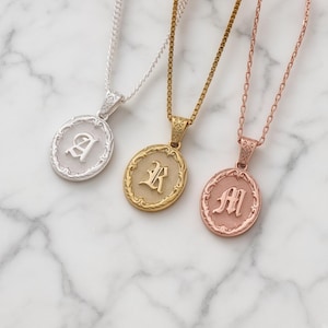 May include: Three oval pendant necklaces featuring the letters A, R, and M in an old English font. The pendants have an ornate, textured border and are suspended from matching metal chains in silver, gold, and rose gold tones respectively.