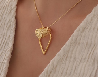 May include: A gold-toned necklace featuring a delicate box chain and a pendant composed of a signet ring with an engraved letter and intricate scrollwork, suspended from a smooth, pointed, V-shaped geometric metal frame.