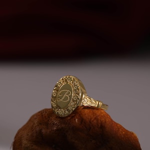 Peut inclure: Une bague chevalière en or présentant une face ovale avec une lettre B cursive gravée au centre. La face ovale est entourée d'un motif floral détaillé, et l'anneau de la bague affiche également des motifs décoratifs gravés en forme de feuilles.