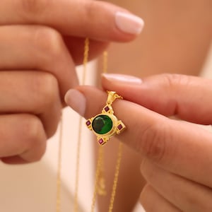 Op de afbeelding: Een gouden hangerketting met een centrale ovaal geslepen groene edelsteen omringd door vier kleine vierkant geslepen rode edelstenen in een kruispatroon. De hanger hangt aan een fijne gouden ketting met een gepolijste metalen afwerking.