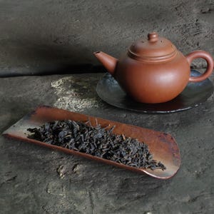 May include: A brown ceramic teapot with a small handle and spout sits on a black saucer. A wooden scoop holds a pile of dark tea leaves. The items are arranged on a textured, gray surface.