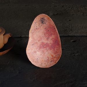 May include: A handcrafted, copper-colored, teardrop-shaped dish with a textured surface. The dish has a reddish-brown hue with darker spots, and is likely used for serving or display. The dish is next to a brown teapot.