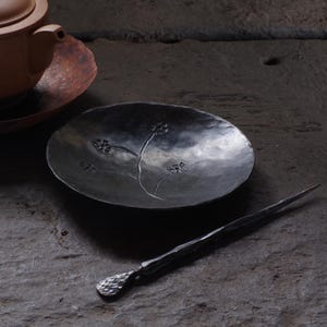 May include: A dark gray, oval-shaped metal dish with a floral design and a long, pointed metal tool. The dish has a hammered texture and the floral design is etched into the surface. The tool has a textured handle.