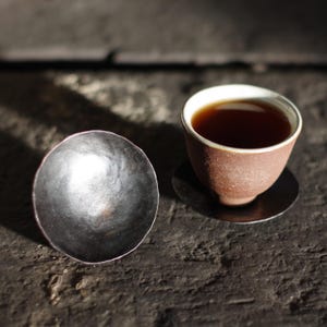 May include: A close-up of a tea set featuring a small, brown ceramic teacup filled with dark tea, resting on a black coaster. Beside it is a small, silver-colored bowl, both set on a dark wooden surface.