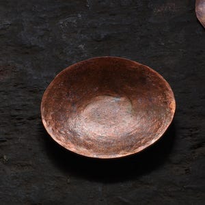 May include: A small, hammered copper bowl with a rounded shape. The bowl has a rich, reddish-brown color with variations in tone, suggesting a handcrafted appearance. The interior is smooth, while the exterior shows a textured surface. The bowl sits on a dark, textured surface.