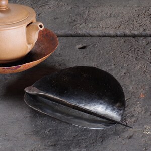 May include: A black, handcrafted tea scoop rests on a dark surface, with a matching tea pot and saucer in the background. The scoop has a curved, leaf-like shape with a long, slender handle. The tea set is made of ceramic.
