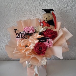 Könnte beinhalten: Ein Blumenarrangement zum Thema Abschlussfeier mit einem Teddybär mit Abschlusskappe und -robe, umgeben von Stoffblumen in Rosa, Rot und Pfirsich. Das Arrangement enthält einen 'Class of 2025'-Topper und ist in pfirsichfarbenes Papier gewickelt.