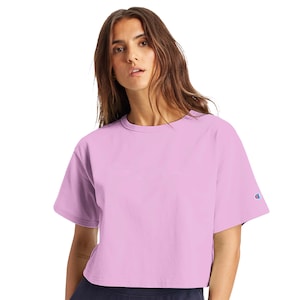 May include: A light purple Champion crop top with short sleeves. The shirt has a crew neck and a small Champion logo on the left sleeve. The model is wearing the shirt.