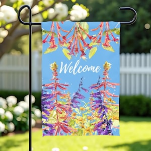 May include: A garden flag with a blue background and the word "Welcome" in white script. The flag features watercolor-style floral designs in red, purple, and yellow. The flag is displayed on a black metal stand in a garden setting.