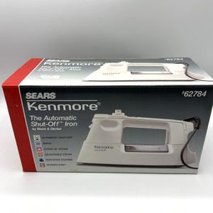 Puede incluir: Una plancha Kenmore blanca con apagado automático en su caja original. La caja es roja, negra y blanca, con el nombre de la marca y las características del producto. La plancha tiene un cable negro y un revestimiento antiadherente.