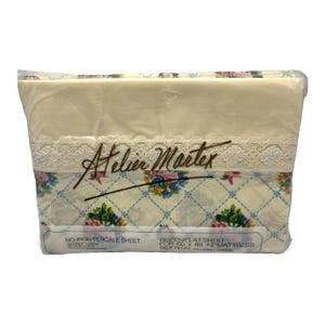 Może przedstawiać: Zapakowane prześcieradło queen firmy Atelier Martex. Prześcieradło jest w kolorze kremowym z kwiatowym wzorem w kolorze niebieskim, różowym i zielonym. Górna krawędź posiada białe koronkowe wykończenie. Opakowanie zawiera tekst "NO-IRON PERCALE SHEET".