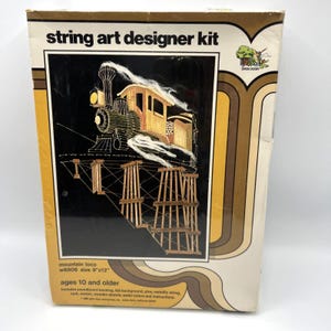 May include: A rectangular string art designer kit box featuring a graphic of a steam locomotive on a trestle bridge. The box is white and yellow with brown wavy lines, labeled string art designer kit, mountain loco, and size 9 inches by 12 inches.