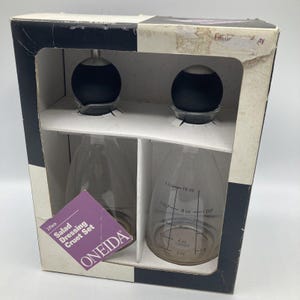 Puede incluir: Un juego de dos jarras para aderezo para ensaladas en una caja. Las jarras son de vidrio transparente con tapas negras y medidas en onzas. La caja es blanca con bordes negros y el logotipo de Oneida. La caja dice "Juego de 2 jarras para aderezo para ensaladas".