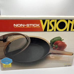 May include: A Vision non-stick covered skillet in a box. The box features the text NON-STICK VERSATILE COOKWARE BY CORNING IT'S VISIBLY SUPERIOR and VISION. The skillet has a dark brown glass lid and handle with a black interior, 10 inches/26 cm.