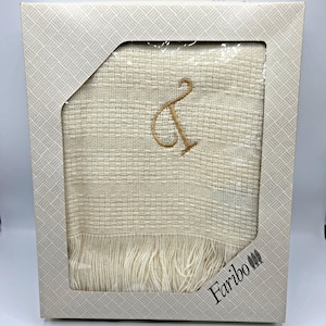 Pode incluir: Cobertor tecido creme com uma letra "&" dourada bordada em uma caixa decorativa. O cobertor tem uma borda com franjas e é rotulado "Faribo".