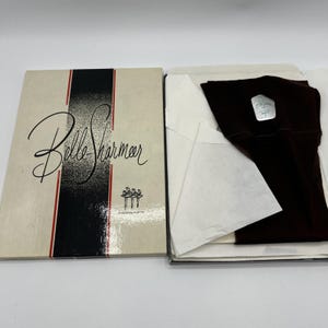 May include: A pair of brown stockings in a white tissue paper-lined box. The box is white with a black and red stripe and the words "Belle Sharmeer" in cursive script. The box also features a small image of three ballerinas.