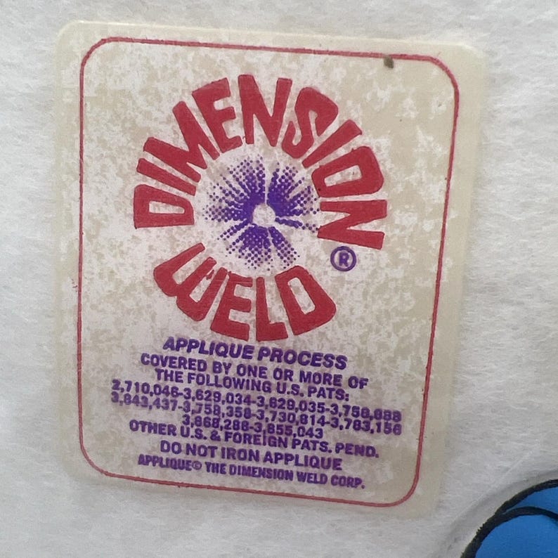 Pu&ograve; includere: Un'etichetta rettangolare con le parole "DIMENSION WELD" in rosso, con un disegno floreale viola e bianco al centro. L'etichetta include anche testo sul processo di applicazione e numeri di brevetto.
