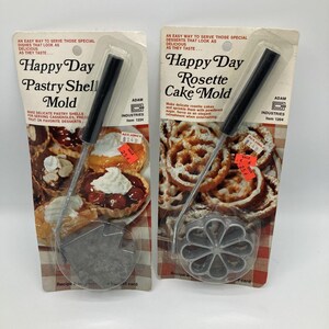 May include: Two vintage Happy Day pastry shell and rosette cake molds. The pastry shell mold is a gray, star-shaped design. The rosette cake mold is a flower-shaped design. Both have black handles and are packaged on a retro-style card.