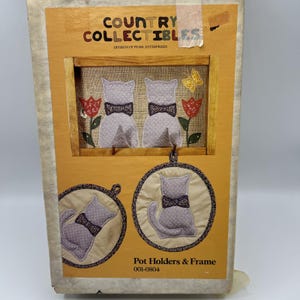 May include: A Country Collectibles box set featuring a framed cat design and two matching pot holders. The frame has two gray cat figures with bow ties, red tulips, and a yellow butterfly. The pot holders have a gray cat design.