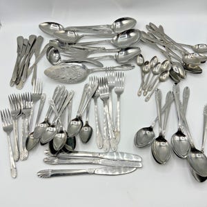 Puede incluir: Una colección de cubiertos plateados vintage, que incluye cucharas, tenedores, cuchillos y utensilios para servir. La plata de ley está dispuesta sobre una superficie blanca, mostrando varios diseños y tamaños. Los utensilios son reflectantes.