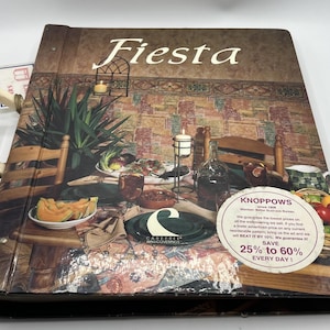 Puede incluir: Una carpeta con la palabra "Fiesta" en escritura blanca. La portada presenta una imagen detallada de una mesa de comedor preparada para una comida, con platos, vasos y comida. Una pegatina en la portada anuncia ahorros.