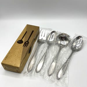 Puede incluir: Un juego de utensilios de servir plateados, que incluye un tenedor, una cuchara, un cucharón y una cuchara de servir, están empaquetados en plástico transparente. Los utensilios tienen un borde decorativo con cuentas. Una caja de cartón marrón con siluetas de utensilios está a la izquierda.