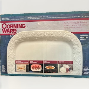 Puede incluir: Una fuente rectangular blanca CorningWare de la colección Vineyard, con un borde en relieve de hojas de vid. El empaque muestra el texto CorningWare, Vineyard y dimensiones de 29,8 cm x 38,1 cm, con imágenes que muestran su uso en microondas, parrilla, horno y lavavajillas.