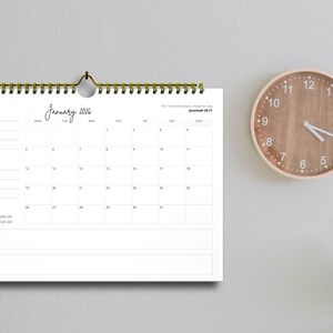 Peut inclure: Un calendrier mural blanc de 2026 avec une reliure à spirale, accroché au mur. Le calendrier affiche le mois de janvier, avec une police manuscrite. Une horloge en bois avec des chiffres noirs est à droite.