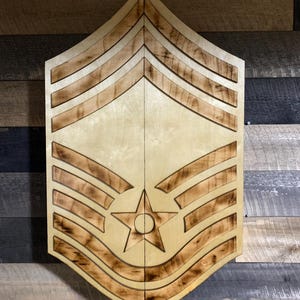 May include: Wooden military rank insignia, featuring a star and stripes design. The light-colored wood is accented with darker, burnt-in lines, creating a layered effect. The piece is mounted on a rustic, multi-toned wooden background.