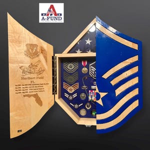 May include: A wooden shadow box with a blue and wood-toned Air Force insignia on the right side. The left side is open, revealing a display of military medals, patches, and pins. The box is engraved with the text "Hurlburt Field FL" and a list of dates and locations.