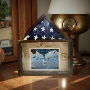 May include: A wooden shadow box with a folded American flag inside. The box also contains a certificate of authenticity with the text "Certificate of Authenticity This Flag was Retired for Patrick Thompson".