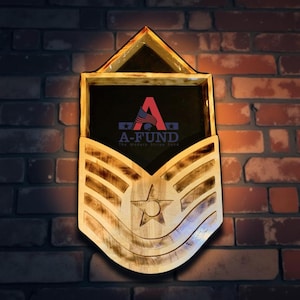 Peut inclure: Décoration murale en bois en forme d'insigne de grade militaire. Le cadre en bois clair entoure un intérieur sombre. L'insigne comporte une étoile et des rayures, avec le texte "A-FUND The Modern Stripe Fund" au centre.