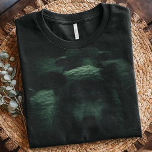 以下が含まれることがあります： 黒地に、黒曜石のラグーンの生き物の薄緑色のグラフィックプリントが施されたTシャツです。