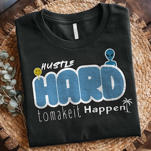 Könnte beinhalten: Schwarzes T-Shirt mit dem Schriftzug "Hustle Hard to make it Happen" in weißen und blauen Buchstaben. Ein gelbes Smiley-Gesicht und eine blaue Alien-Grafik sind ebenfalls abgebildet. Das T-Shirt liegt auf einer gewebten Oberfläche.