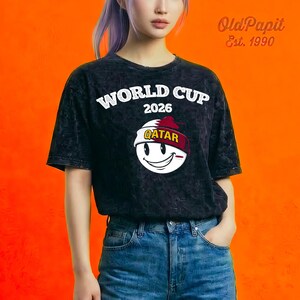 May include: Black, acid-wash t-shirt with the words "WORLD CUP 2026" in white. Below the text is a smiling face wearing a headband that says "QATAR". The shirt has short sleeves and a crew neck.