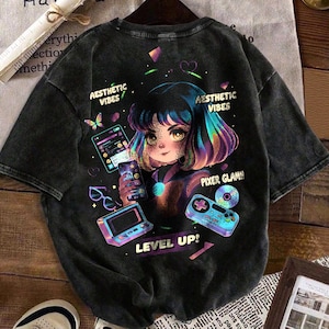 May include: Black t-shirt with a vibrant anime-style graphic. The design showcases a girl with rainbow hair, holding a phone and gaming accessories. Text includes "Aesthetic Vibes," "Pixer Glam!," and "Level Up!"