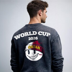 May include: Dark gray sweatshirt with the text "WORLD CUP 2026" in white. Below the text is a smiley face design with the word "QATAR" on a red and white headband. The sweatshirt is a casual style.
