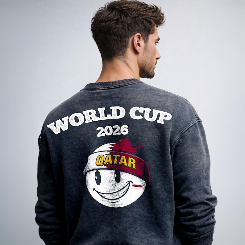 May include: Dark gray sweatshirt with the text "WORLD CUP 2026" in white. Below the text is a smiley face design with the word "QATAR" on a red and white headband. The sweatshirt is a casual style.